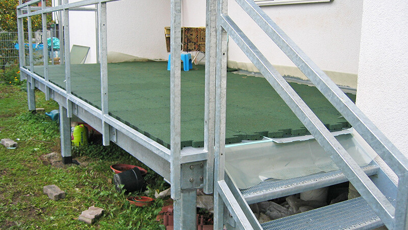 Aus Metall gefertigte Terrasse mit grünen Platten, die an ein Haus angrenzt. Eine kleine Treppe führt zwei Stufen hinauf. Der Unterbau besteht aus Gitterrost.