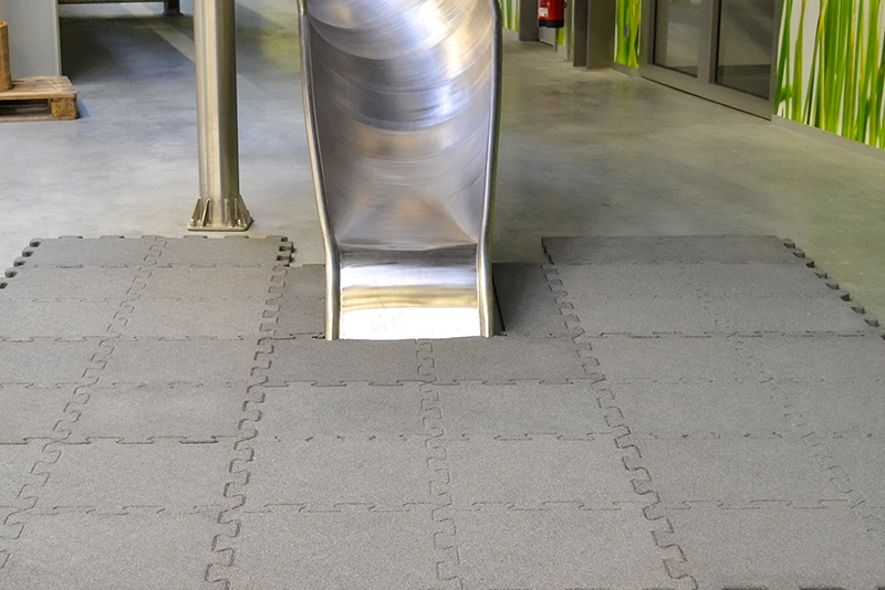 Das Bild zeigt den unteren Teil einer Edelstahlrutsche, die in einem Indoorbereich installiert wurde. Vor der Rutsche wurden graue Fallschutzplatten von WARCO verlegt, die durch eine Puzzleverzahnung an den Kanten formschlüssig und stabil miteinander verbunden sind. Diese robusten und langlebigen Fallschutzplatten bieten eine sichere und stoßdämpfende Oberfläche, um Verletzungen bei Stürzen zu vermeiden. Die Platten eignen sich hervorragend für Indoor-Spielplätze oder Kletterhallen, in denen Sturzgefahr besteht. Im Hintergrund sind die Wände mit einem natürlichen, grasgrünen Dekor versehen, das dem Raum eine angenehme Atmosphäre verleiht.