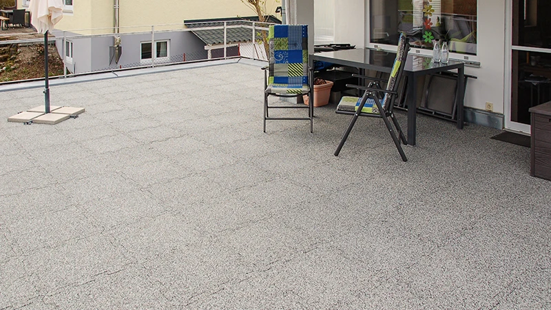 Auf einer Dachterrasse liegen grau marmorierte WARCO-Terrassenplatten aus pu-gebundenem Gummigranulat. Ihre Puzzleverzahnung sorgt für eine passgenaue Verbindung und vereint Witterungsbeständigkeit mit Langlebigkeit. Das formstabile Material dämpft Trittgeräusche und trägt zu einem aufgeräumten, gepflegten Erscheinungsbild bei. Zudem bietet es Rutschhemmung und hohen Gehkomfort, was den Belag besonders nutzerfreundlich macht.