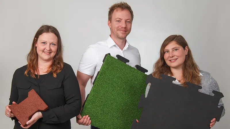 Fachberatung für langlebigen Balkonboden Lächelnd präsentieren drei Fachberater von WARCO verschiedene Bodenplatten aus pu-gebundenem Gummigranulat in Braun, Grün und Schwarz. Diese Platten eignen sich besonders für Balkone und bieten rutschhemmende, langlebige Eigenschaften. Jede Oberfläche hat ein eigenes Design und lässt sich einfach verlegen. Mit ihrer kompetenten Beratung unterstützen die Experten bei der Wahl des passenden Bodenbelags, damit jeder Balkon sicher und ansprechend gestaltet werden kann. Die pflegeleichten Platten überzeugen durch Strapazierfähigkeit und Komfort.