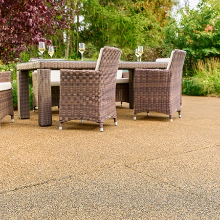 Blick auf eine sonnige Terrasse mit beigebraunen Terrassenplatten von WARCO. Der Bodenbelag aus pu-gebundenem Gummigranulat ist rutschfest, wasserdurchlässig und sorgt für hohen Gehkomfort. Ein stabiler Gartentisch mit geflochtenen Stühlen und gefüllten Weingläsern lädt zum Entspannen ein. Rundherum blühen üppige Sträucher und bunte Blumen, die eine naturnahe Atmosphäre schaffen. Die elastische Oberfläche ist frostbeständig, dämpft Trittgeräusche und bleibt formstabil.
