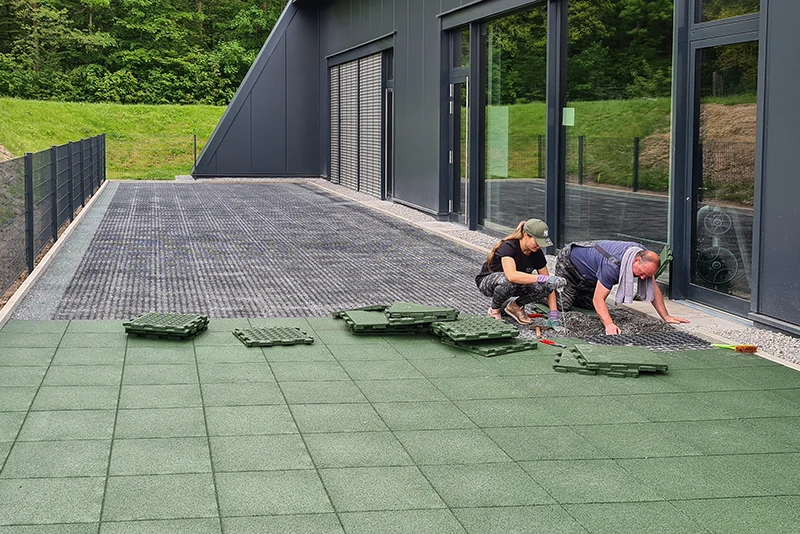 Vor einer dunklen, modernen Fassade verlegen eine Frau und ein Mann grüne WARCO-Terrassenplatten aus pu-gebundenem Gummigranulat. Die Platten werden passgenau aneinandergereiht, um eine rutschfeste, witterungsbeständige und langlebige Oberfläche auf der Terrasse zu schaffen. Dieses robuste Material bietet Komfort und Schutz bei jedem Wetter. Die einfache Verlegung ermöglicht eine individuelle Gestaltung und verleiht jeder Terrasse ein hochwertiges, zeitgemäßes Erscheinungsbild.