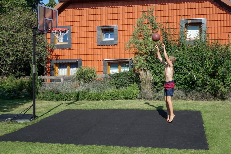Im Garten bei einem Einfamilienhaus liegt ein Basketballfeld aus schwarzen Ballspielplatten von WARCO. Ein Junge wirft den Basketball auf den Basketballkorb, der mobil aufgestellt wurde.