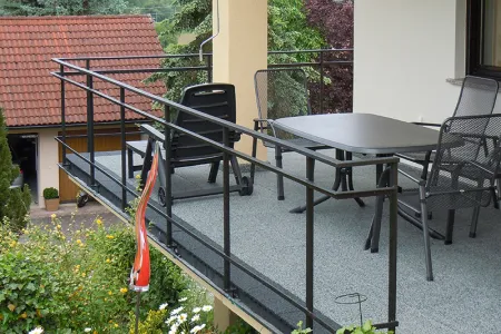 Auf dem Bild sieht man einen geräumigen Balkon, dessen Boden mit grauen WARCO-Bodenplatten ausgelegt ist. Ein schwarzes Metallgeländer umrahmt die Fläche und bietet Sicherheit. Dazu passen der großzügige Tisch und die Stühle, die zum geselligen Beisammensein einladen. Der elastische, rutschfeste Belag ist witterungsbeständig und pflegeleicht, wodurch dieser Außenbereich dauerhaft geschützt ist und zum Entspannen einlädt.
