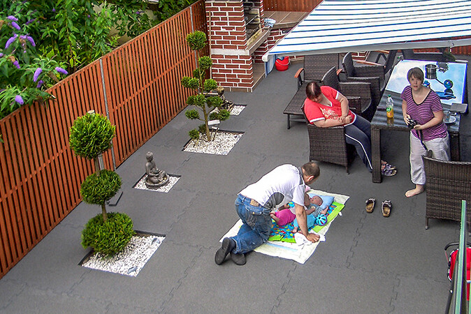 Eine Familie genießt ihre freie Zeit auf einer als Wohlfühl-Oase gestalteten Terrasse mit grauen Terrassenbelag von WARCO. Das Baby liegt auf eine Kinderdecke auf den Terrassenplatten